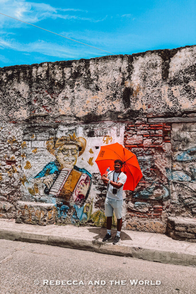 A person - our guide on a free Cartagena walking tour - holding a bright orange umbrella stands beside a mural of a musician painted on an old stone wall, a street-art scene for what to do in Cartagena.