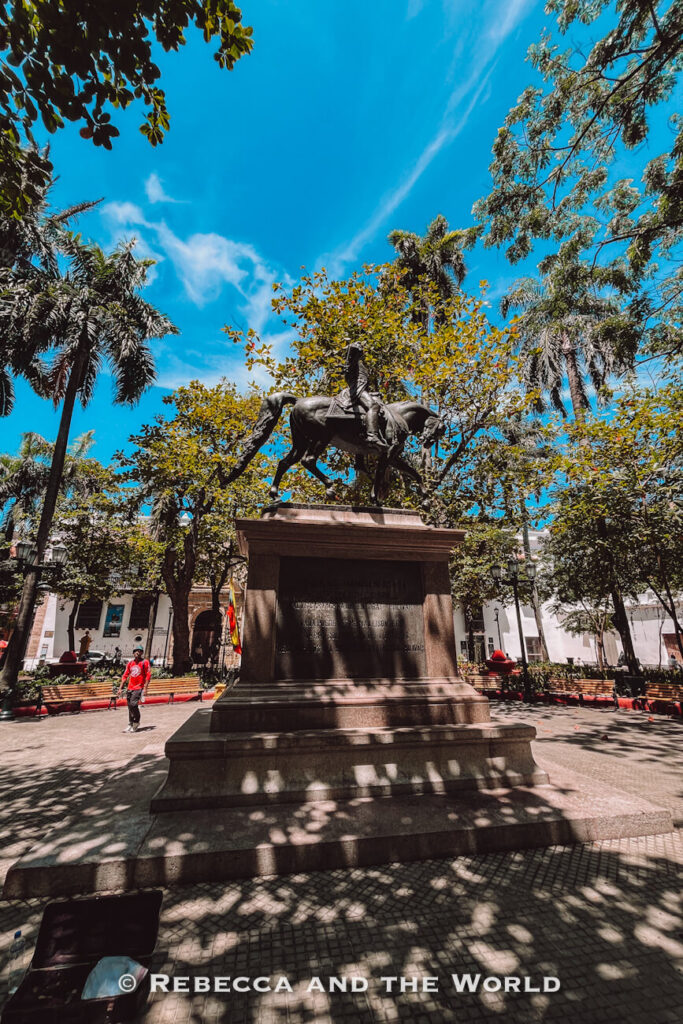 A statue of Simon Bolivar stands on a large stone pedestal in a leafy plaza shaded by tall trees, a historic stop on a Cartagena itinerary.