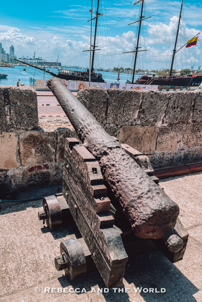 An old iron cannon points over a stone fortification wall toward the harbour, where tall sailing ships are docked, a landmark in this Cartagena travel guide.