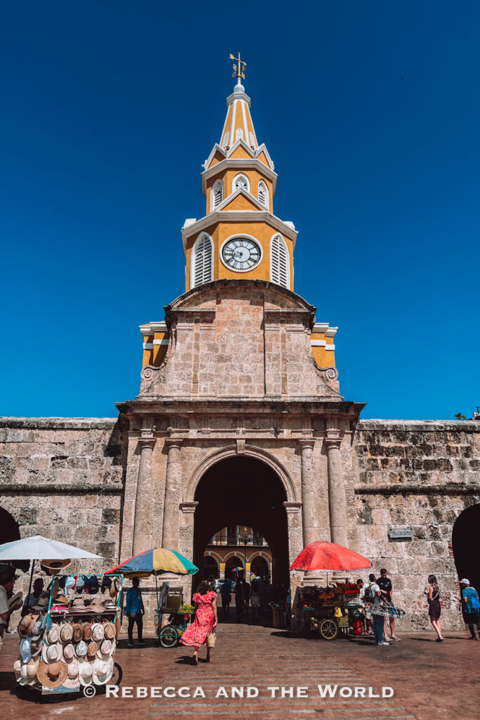 A stone gateway with a clock tower rises above a busy entrance lined with market stalls and visitors, one of the classic things to do in Cartagena.