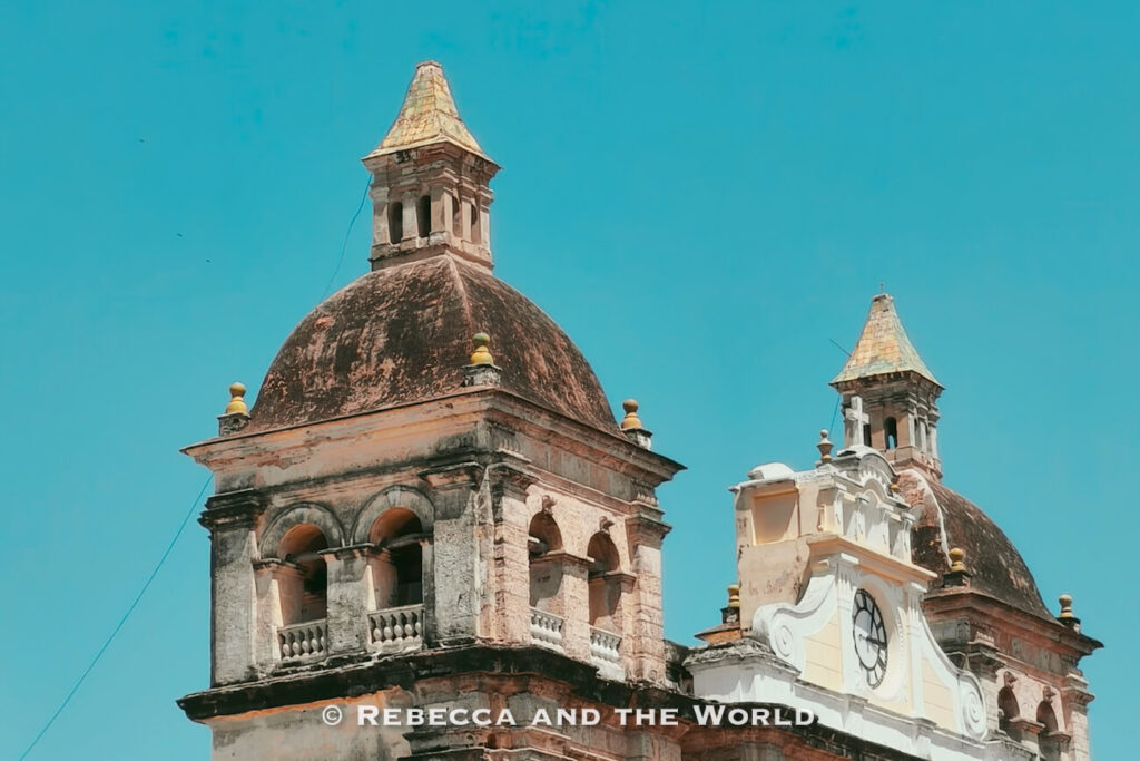 Two weathered church bell towers with domed roofs rise against a bright blue sky, with a clock visible on the façade below. Visiting historic churches is among the top things to do in Cartagena.