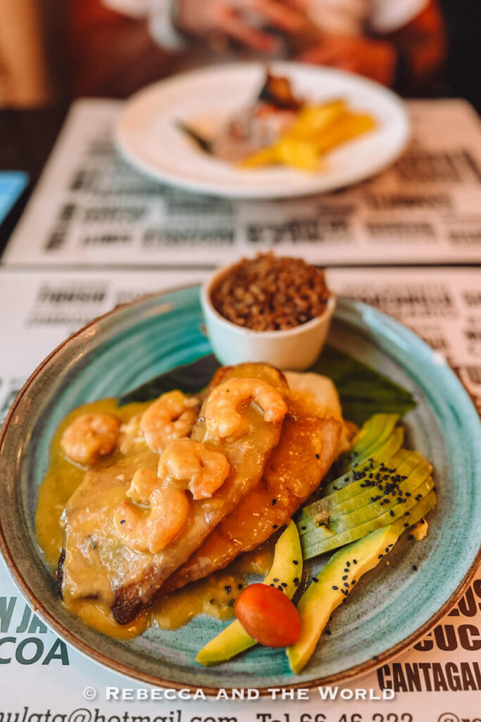 A plate of fish topped with shrimp in a rich sauce is served with sliced avocado, a cherry tomato and a side of rice, highlighting local cuisine in this Cartagena travel guide.