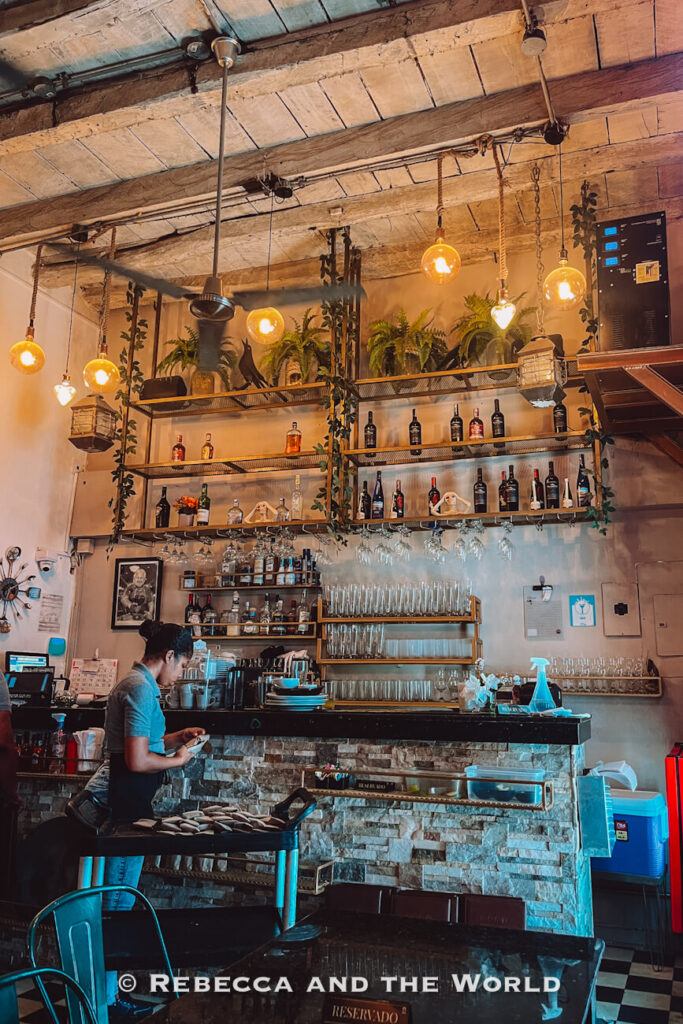 A restaurant - called La Mulata - interior features hanging globe lights, exposed wooden ceiling beams, trailing plants and shelves lined with wine bottles above the bar, one of the atmospheric things to do in Cartagena.
