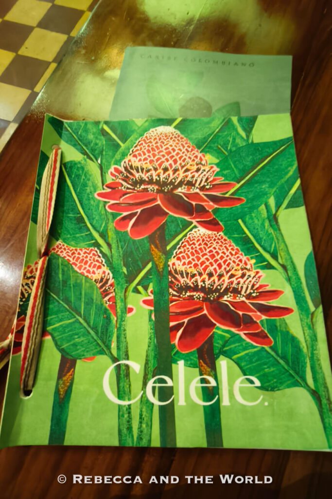 A colorful restaurant menu featuring large tropical flowers rests on a wooden table, with a woven bookmark placed along the left edge. The menu highlights local flavors featured in many Cartagena travel guide recommendations. Celele is one of the best restaurants in Cartagena.