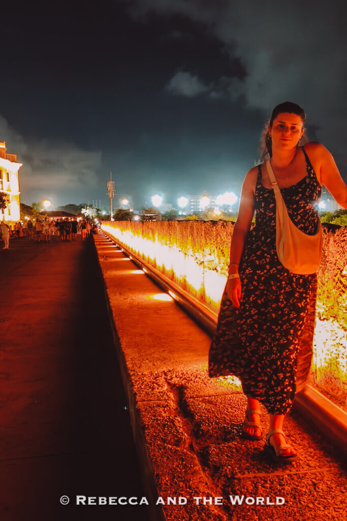 A woman (the author of this article)wearing a dark patterned dress and white crossbody bag stands on a stone walkway beside an illuminated historic wall at night. Walking the city walls after dark is a highlight of 3 days in Cartagena.