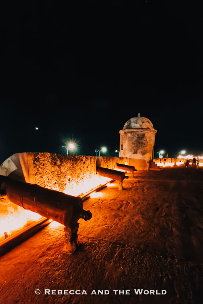 Historic cannons line a stone fortification wall lit with warm orange lights, while a small guard tower stands under the night sky. Exploring Cartagena’s fortifications is a classic thing to do in Cartagena.
