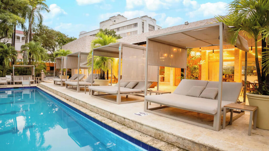 White poolside cabanas line the edge of a turquoise swimming pool surrounded by palm trees and tropical landscaping, a relaxing option for where to stay in Cartagena.