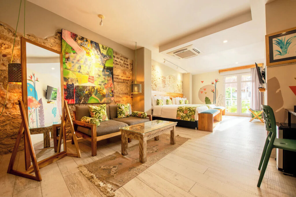 A spacious hotel room features exposed stone walls, tropical-inspired artwork, a seating area and two large beds, offering comfortable accommodation for 3 days in Cartagena.