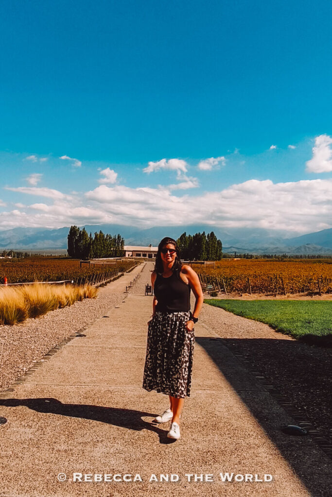 A woman (the author of this article) wearing sunglasses, a black sleeveless top, a patterned midi skirt, and white sneakers stands on a wide gravel-and-concrete pathway running through a vineyard in Mendoza. Rows of grapevines stretch out on both sides of the path, with a winery building in the distance and mountains visible beneath a bright blue sky filled with scattered white clouds. This vineyard setting provides inspiration for what to wear in Argentina, especially for wine regions where comfortable footwear and lightweight layers work well.