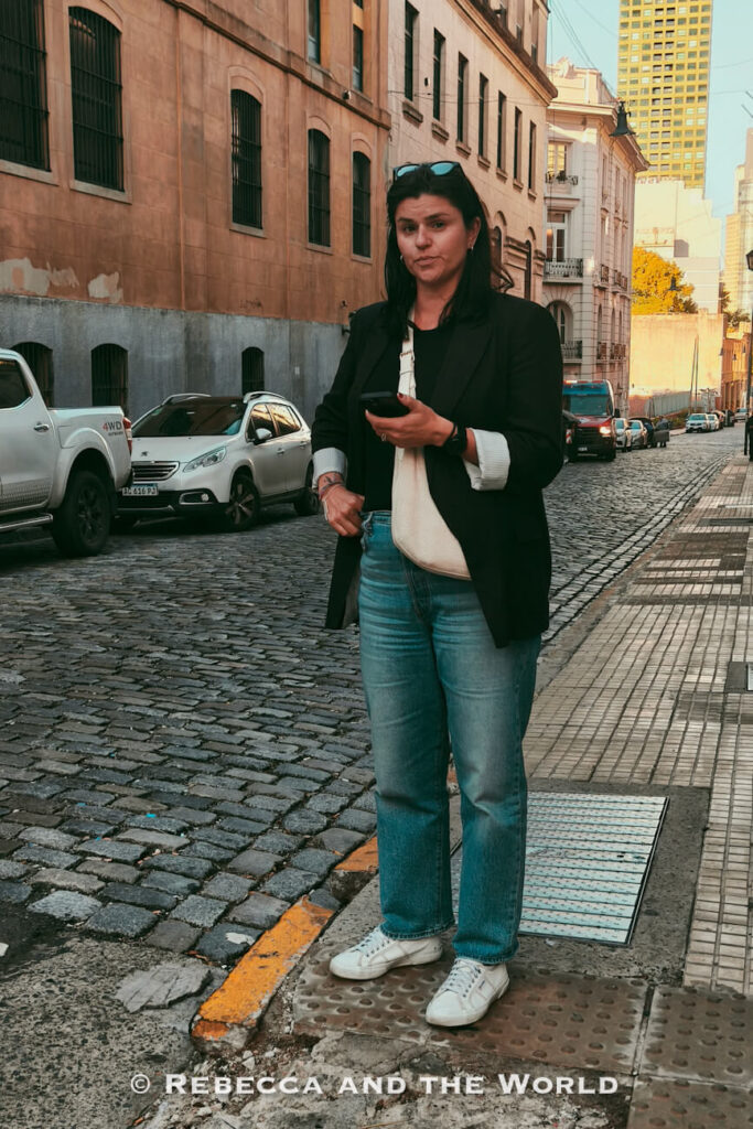 A woman (the author of this article) stands on a cobblestone city street in Buenos Aires lined with historic buildings and parked cars. She is wearing light-wash jeans, white sneakers, a black top, and a dark blazer, while holding a phone and carrying a white crossbody bag. Tall buildings rise in the background, creating a mix of old and modern urban architecture. This outfit is a practical example for an Argentina packing list, combining comfort and style for exploring cities on foot.