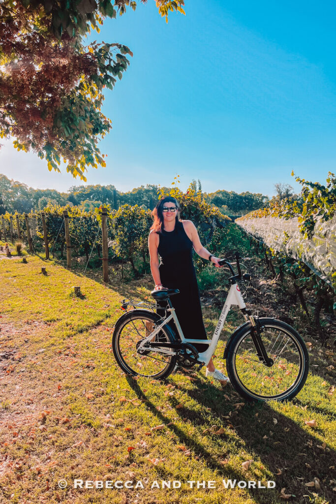 A woman in a sleeveless black dress and white sneakers stands beside a white bicycle in a vineyard in Mendoza. Sunlight filters through nearby trees, casting long shadows across the grassy ground, while rows of grapevines extend into the distance under a clear blue sky. The relaxed outfit and comfortable shoes are useful inspiration when deciding what to pack for Argentina if your itinerary includes wine tours and outdoor activities.