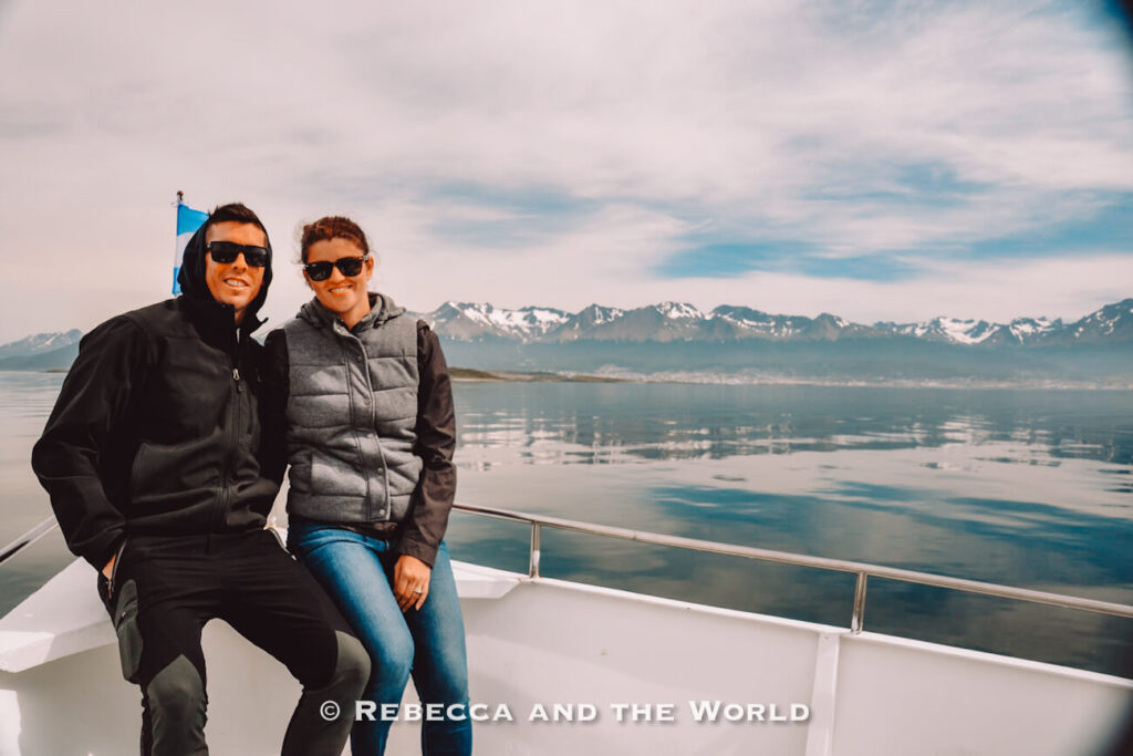 Two people - the author and her husband - wearing sunglasses and warm outerwear sit together on the bow of a boat. Behind them, calm water reflects a mountain range with snow-capped peaks beneath a cloudy sky. The scene conveys a cool-weather adventure in a remote natural setting, highlighting why insulated layers belong on an Argentina packing list for southern regions and boat excursions.