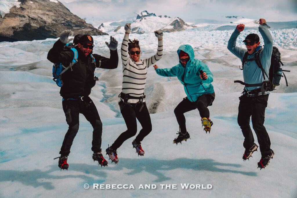 Four people wearing outdoor jackets, gloves, hiking gear, and crampons are captured mid-jump on the surface of a glacier. Snow-covered mountains and icy formations fill the background, emphasizing the rugged alpine environment. Their specialized cold-weather clothing demonstrates what to pack for Argentina when visiting glaciers and undertaking outdoor excursions in Patagonia.
