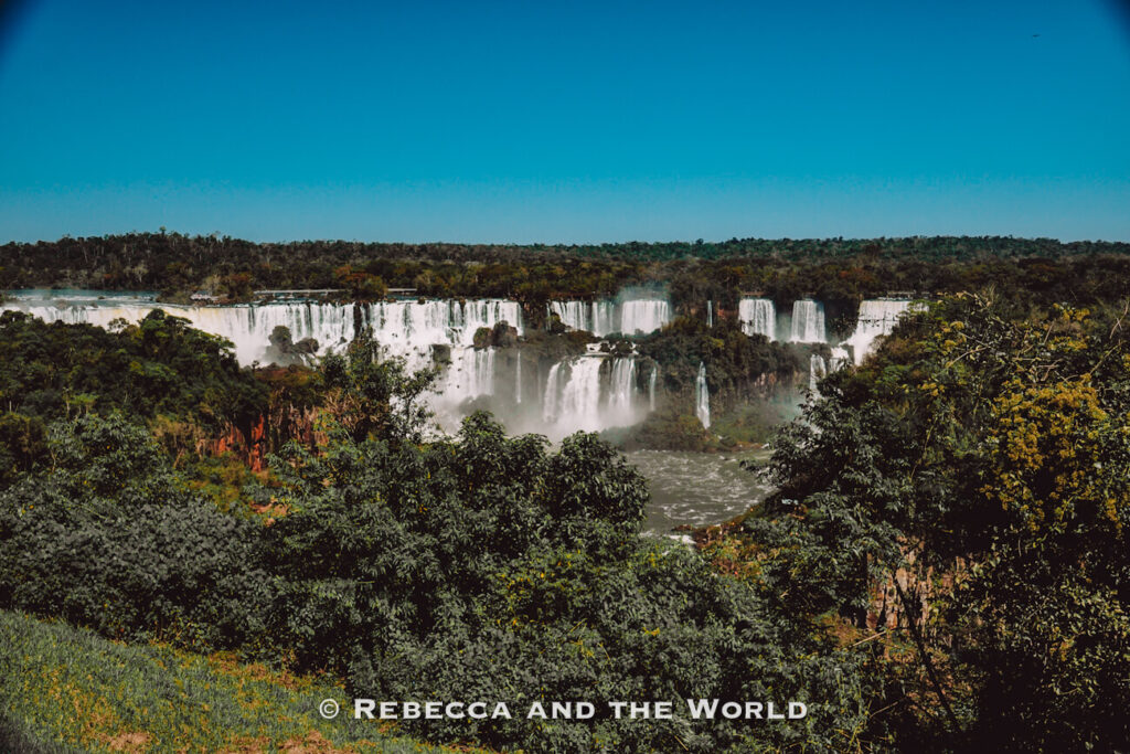 A panoramic landscape view of Iguazu Falls cascading through dense green rainforest. Multiple waterfalls plunge into a river below, with mist rising from the falls and a clear blue sky overhead. The lush vegetation and warm-weather setting illustrate why lightweight clothing, sun protection, and breathable fabrics are important when considering what to wear in Argentina for subtropical destinations.