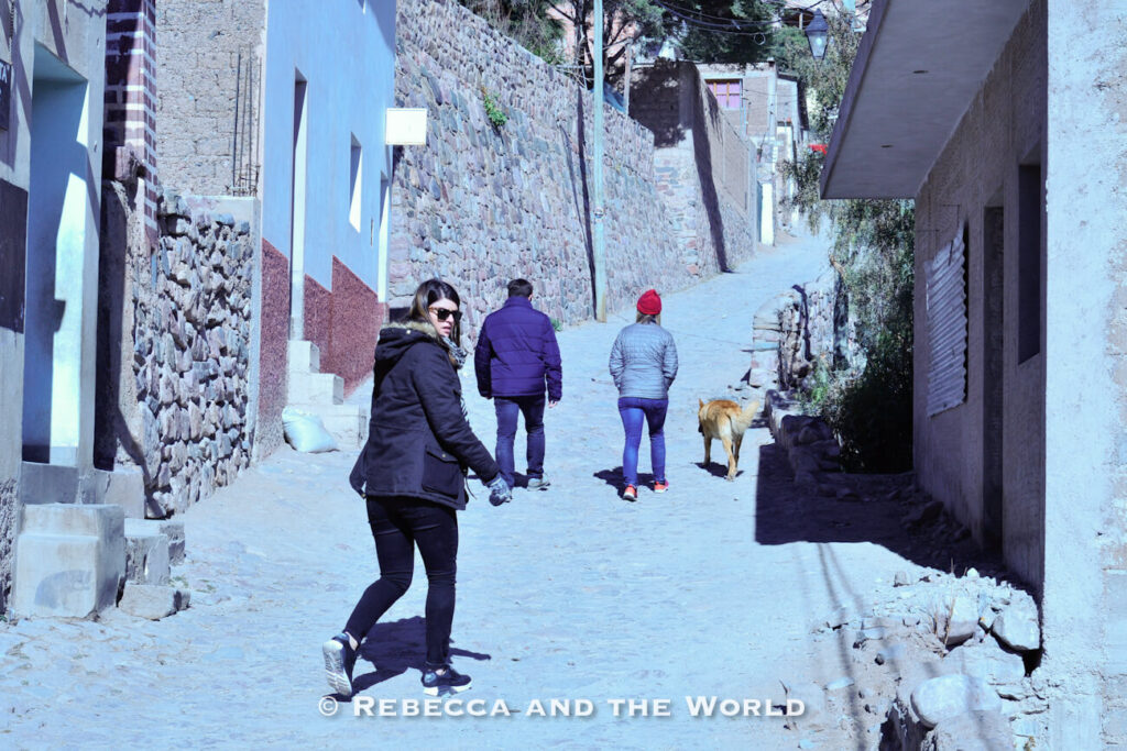 A woman (the author of this article) wearing a black winter jacket, black leggings, gloves, sunglasses, and athletic shoes walks up a narrow cobblestone street in a small hillside town in north Argentina. Stone walls and simple buildings line both sides of the road, while two other pedestrians and a dog continue farther up the steep incline. Bright sunlight creates strong shadows across the street, highlighting the rugged terrain and cool-weather conditions. This practical layered outfit offers useful inspiration for an Argentina packing list, especially when visiting mountain towns where chilly mornings and uneven streets call for warm clothing and comfortable walking shoes.