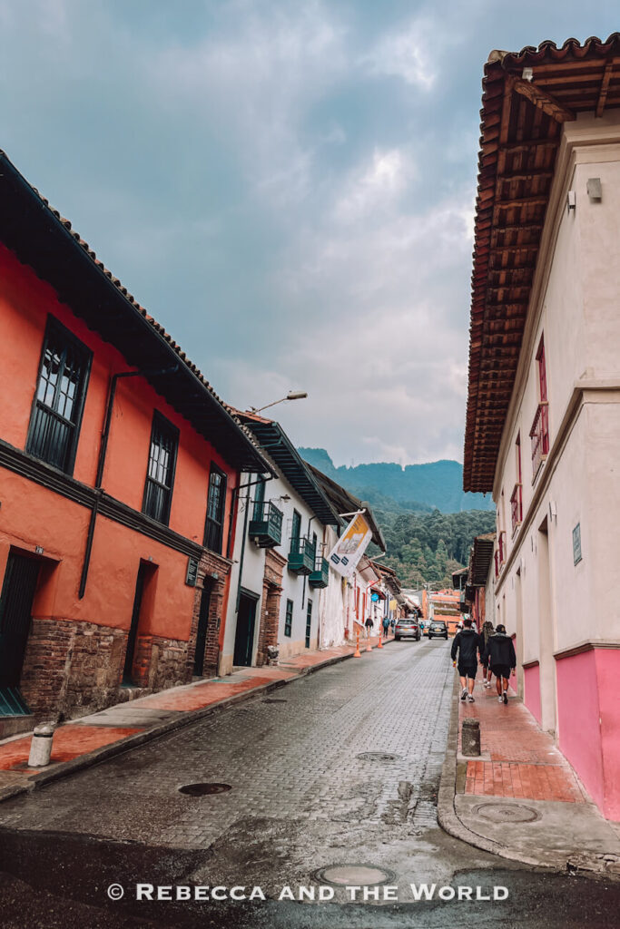 Rainy cobblestone street in La Candelaria, Bogot&aacute; lined with colonial buildings, red sidewalks, tiled roofs, and green mountains in the background during a Bogot&aacute; food tour.