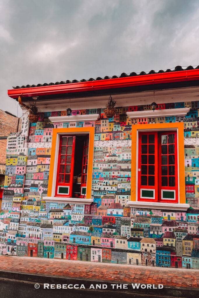Colorful Bogot&aacute; building fa&ccedil;ade covered in a painted mural of tiny houses, with bright red windows and orange trim on a cloudy day during a Bogot&aacute; food tour.