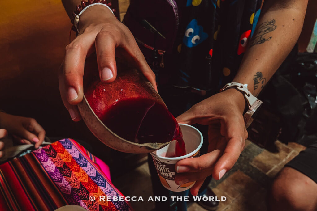 Close-up of a person pouring a dark red drink - chicha - from a clay cup into a small paper cup during a Colombian food tasting.