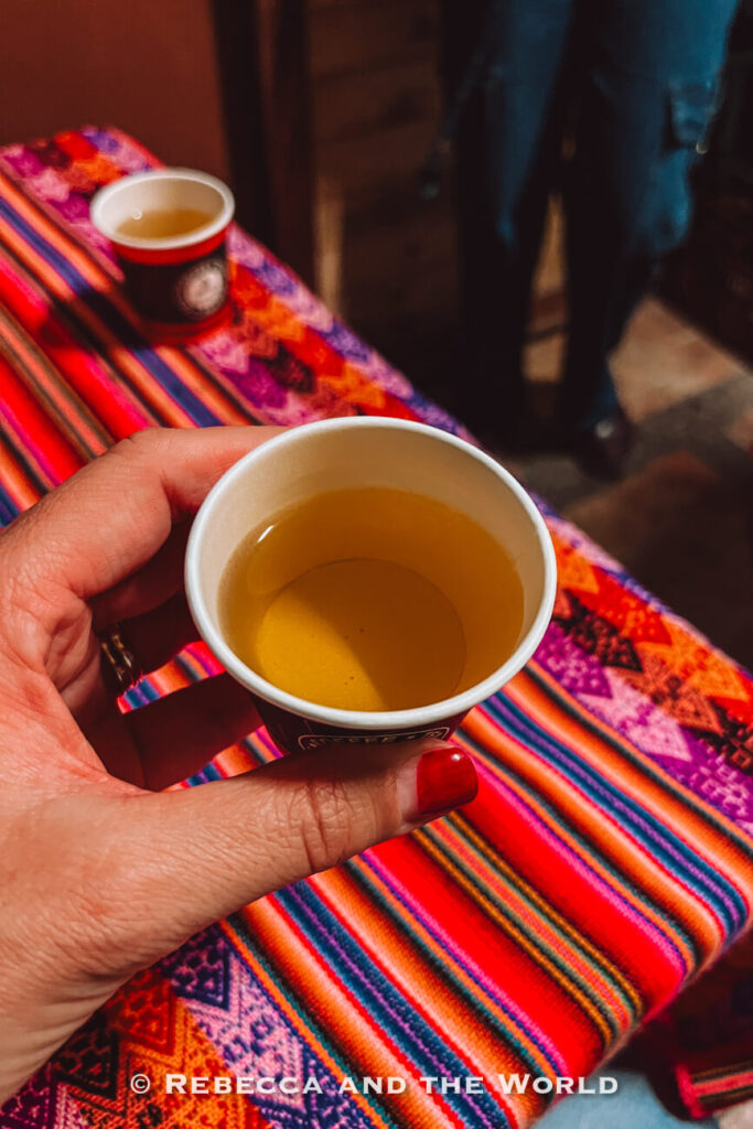 Hand holding a small paper cup of golden herbal tea - coca tea - above a bright woven textile during a Bogot&aacute; food tour.