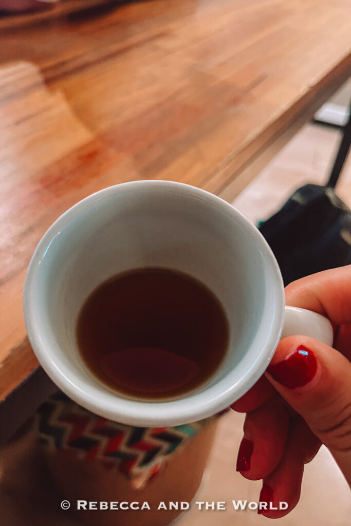 Close-up of a hand holding a small white cup of dark coffee above a wooden table during a Bogot&aacute; food tour.