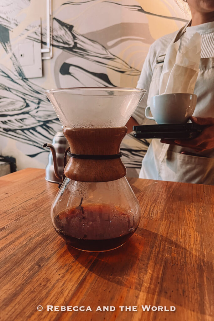 Glass Chemex coffee brewer with dark coffee on a wooden table while a barista holds a cup and filter, showing Colombian food and coffee culture.
