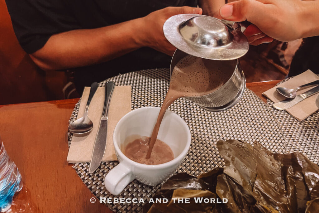 Frothy hot chocolate being poured from a metal pitcher into a white mug on a restaurant table during a traditional Colombian food tasting.