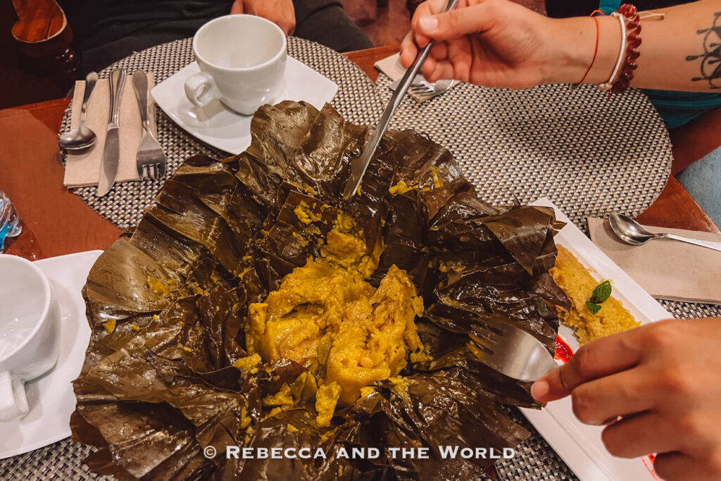 Large banana-leaf-wrapped Colombian dish of tamal opened on a white platter, revealing a yellow filling being shared with forks.
