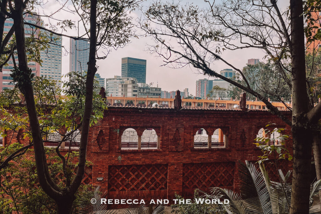 Historic red-brick building with arched windows framed by trees, with modern high-rise buildings in the background during a Bogot&aacute; food tour.