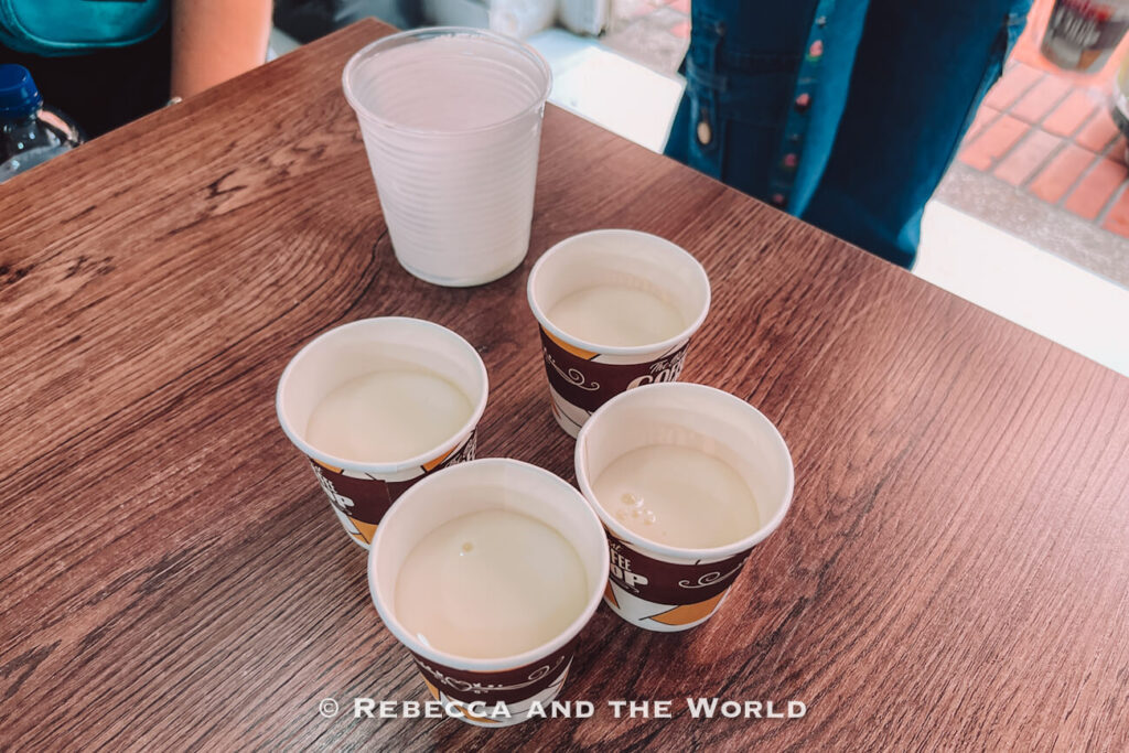 Four small paper cups filled with a pale white drink - oat cream -arranged on a wooden table during a Colombian food tasting.
