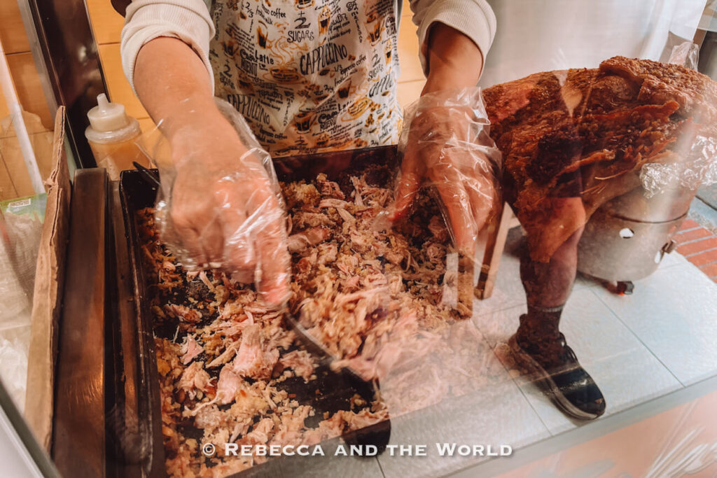Vendor wearing plastic gloves mixes shredded roasted pork behind a glass counter, with a large piece of crispy pork skin nearby for a traditional Colombian food tasting.