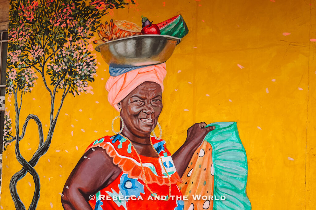 Bright yellow wall mural of a smiling woman in colorful clothing balancing a bowl of fruit on her head during a Bogot&aacute; food tour.