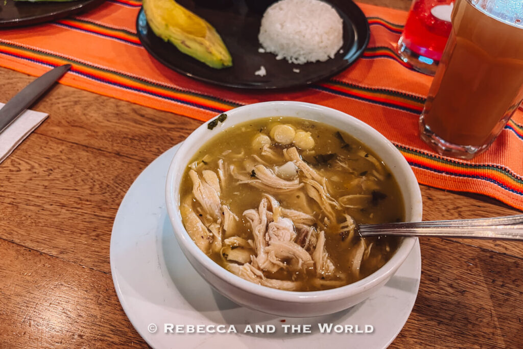 Bowl of chicken soup with shredded chicken, corn, and herbs, served with rice, avocado, and a drink as a traditional Colombian dish. Ajiaco is one of the best Colombian foods to try.
