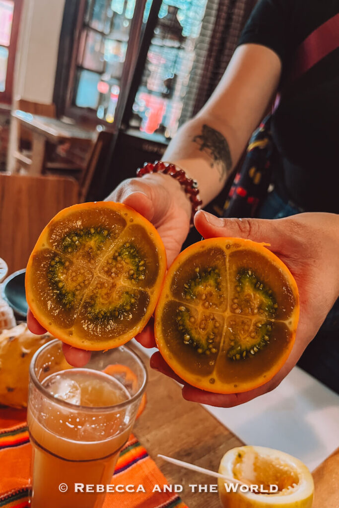Person holding two halves of a yellow-orange tropical fruit with green seeds and jelly-like flesh during a Colombian food tasting.