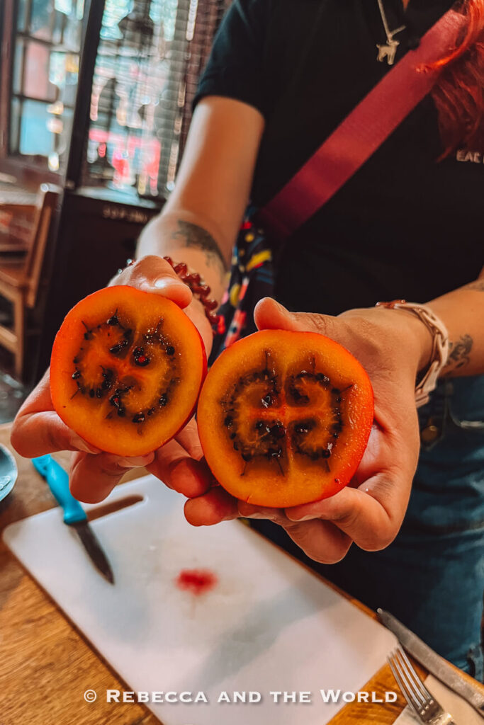 Person holding two halves of a bright orange tropical fruit with dark seeds and soft pulp during a Bogot&aacute; food tour.