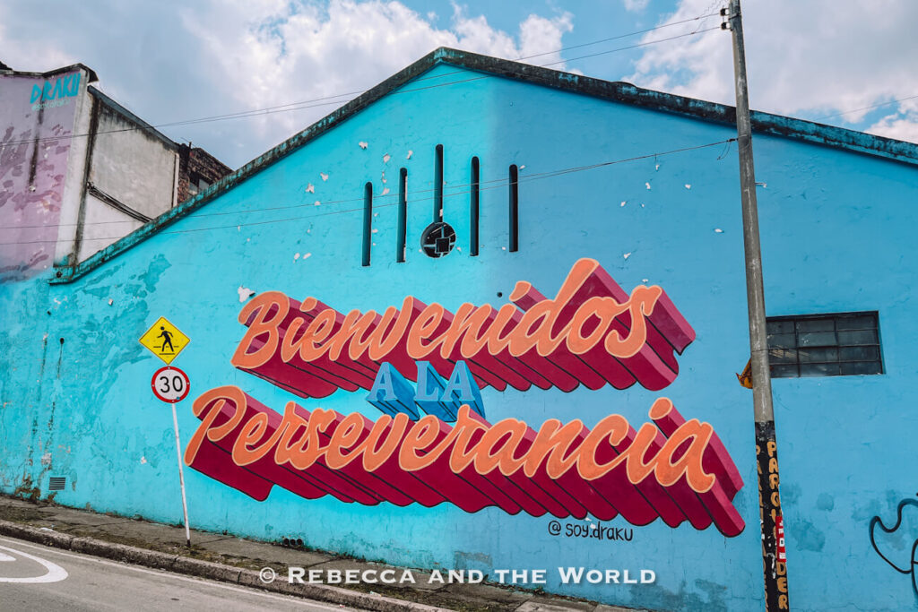 Large turquoise building in Bogot&aacute; featuring a colorful mural that reads &ldquo;Bienvenidos a la Perseverancia&rdquo; in bold pink and orange lettering, with a pedestrian crossing sign, speed limit sign, and cloudy sky overhead during a Bogot&aacute; food tour.