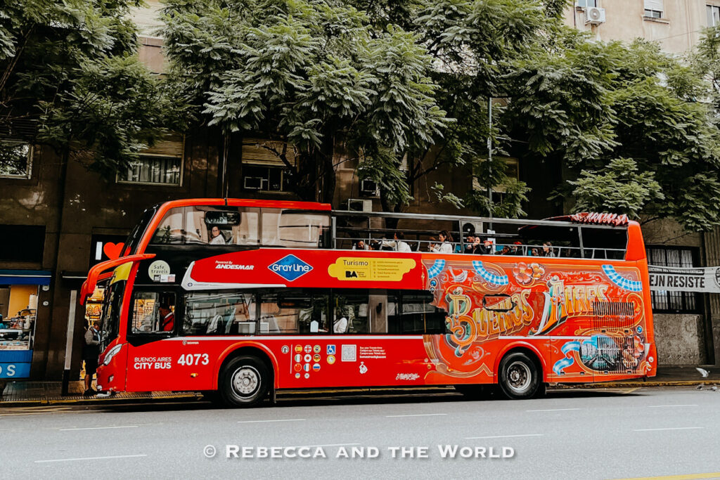A bright red open-top double-decker Buenos Aires hop on hop off bus is parked along a tree-lined city street in Buenos Aires. The Buenos Aires tourist bus features colorful artwork on the side with the words “Buenos Aires” in large decorative lettering. Several passengers are seated on the upper deck while apartment buildings and leafy green trees fill the background. The bus displays tourism logos and accessibility symbols along the lower side panel.