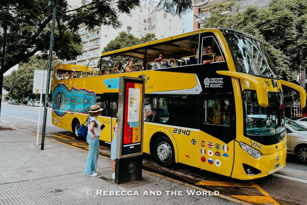 A yellow Buenos Aires tourist bus stopped at a curbside bus stop on a wide city street surrounded by trees and tall residential buildings. The open-top Buenos Aires hop on hop off bus has passengers seated on the upper level, while two women stand on the sidewalk near the boarding area. The side of the bus is decorated with colorful graphic artwork, and international flag icons are displayed near the front wheel.