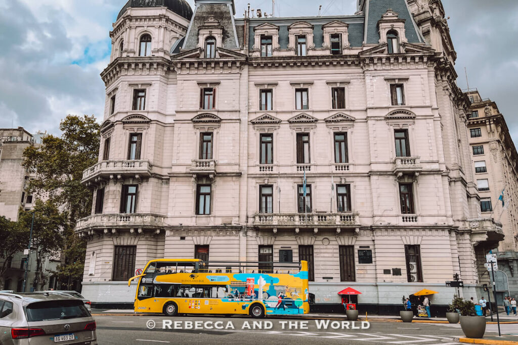 A yellow double-decker Buenos Aires tourist bus drives past a large historic building with ornate architecture and tall windows in downtown Buenos Aires. The open-top bus is decorated with colorful city-themed illustrations, while cars and pedestrians move along the wide street under a cloudy sky.