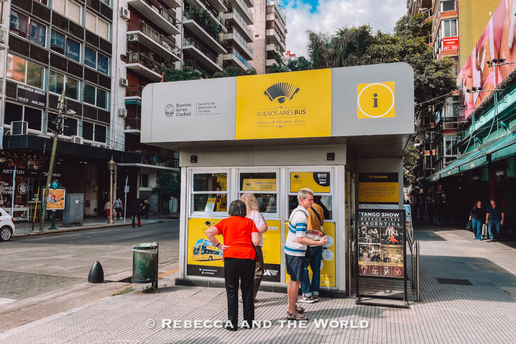 People stand outside a Buenos Aires hop on hop off bus ticket booth on a busy city street lined with tall apartment buildings and storefronts. The small kiosk features yellow and white signage for the Buenos Aires Bus service, with posters and tourist information displayed outside.