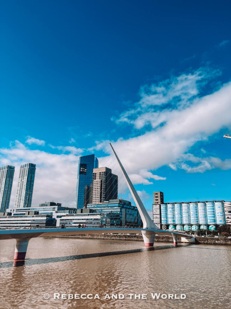 The modern Puente de la Mujer bridge stretches across the water in Puerto Madero, Buenos Aires, with tall glass skyscrapers and apartment buildings in the background. The white pedestrian bridge stands beneath a bright blue sky with large white clouds reflected in the river below.