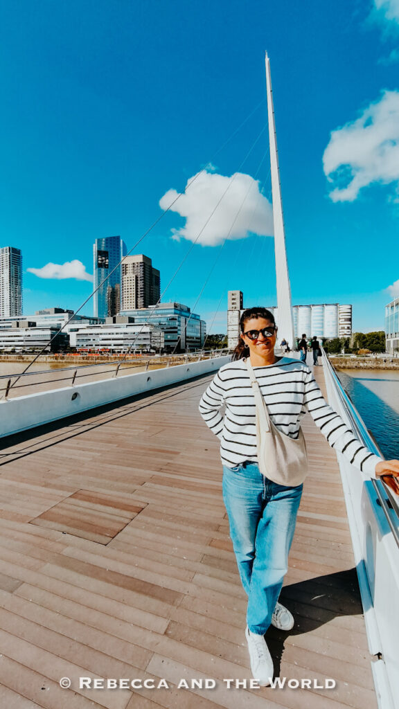 A woman (the author of this article) wearing sunglasses, a striped sweater, jeans, and white sneakers stands smiling on the Puente de la Mujer bridge in Buenos Aires. Modern high-rise buildings and blue skies with scattered clouds form the background along the waterfront walkway.
