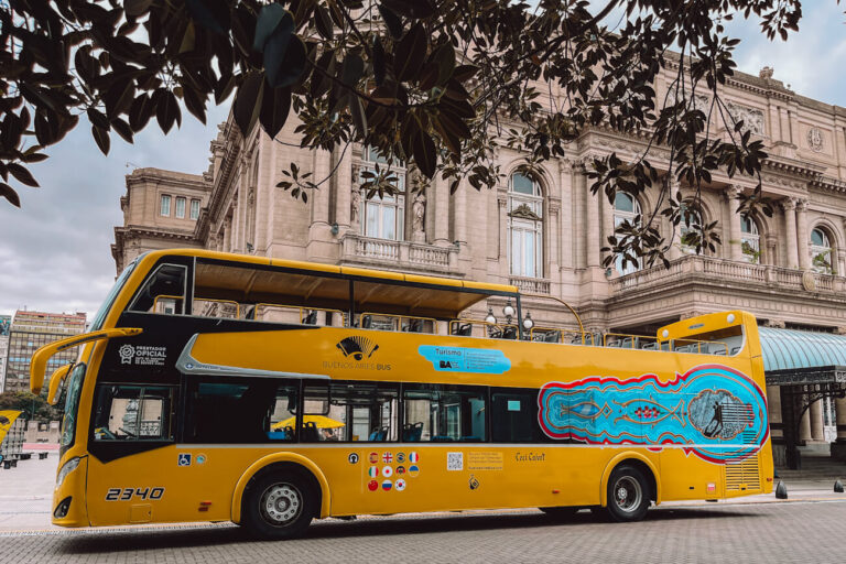 A bright yellow double-decker sightseeing bus is parked in front of an ornate historic building in Buenos Aires. Tree branches frame the top of the image, while colorful artwork decorates the side of the bus. The Buenos Aires hop on hop off bus is a great option for visitors exploring the city.