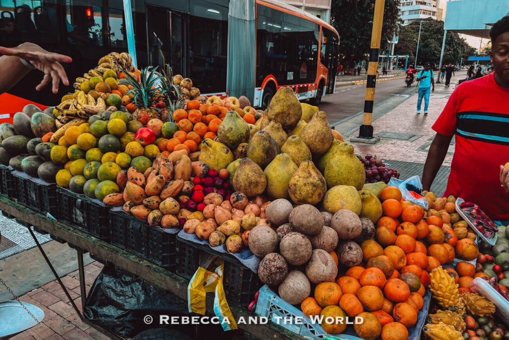A large roadside fruit cart displays heaps of oranges, pears, bananas, coconuts, and tropical fruit beside a city bus stop during a Cartagena food tour.