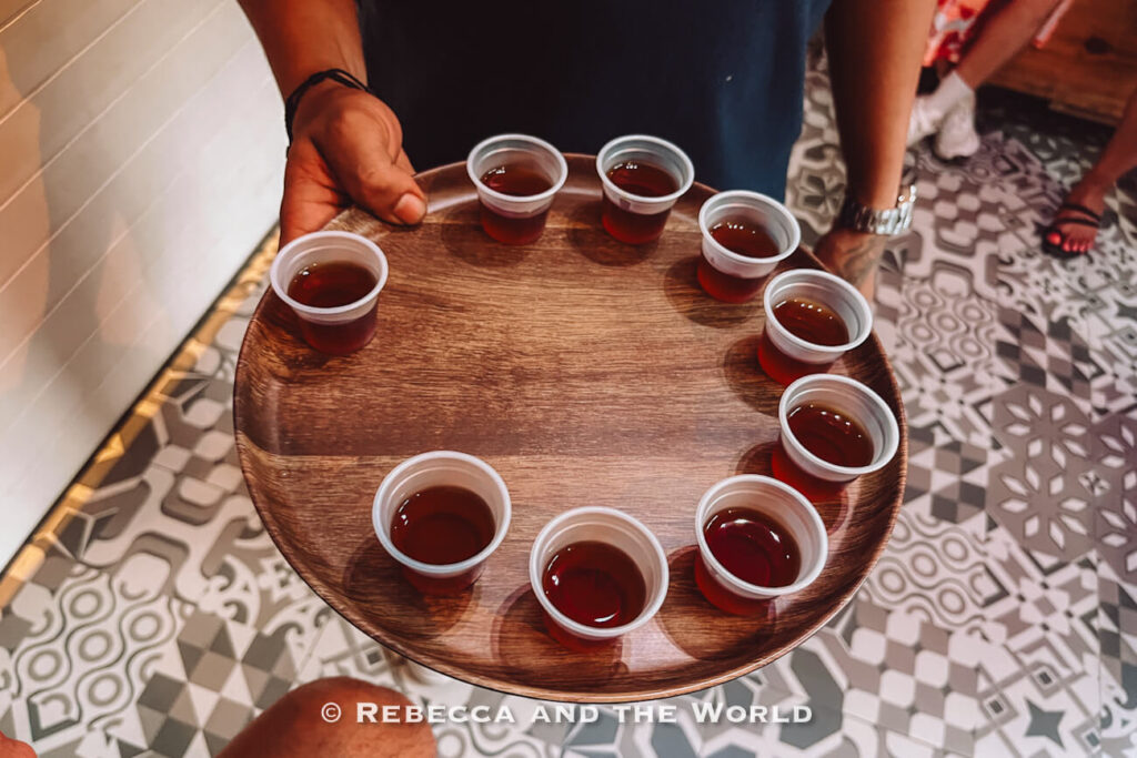 A server carries a wooden tray with small plastic cups filled with a coffee tasting sample inside a restaurant with patterned tile flooring.