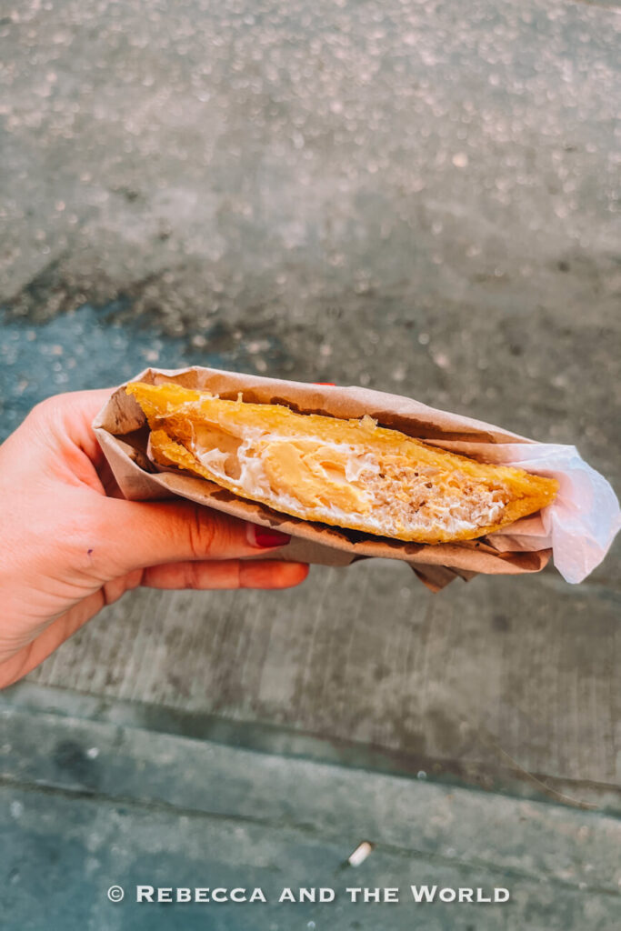 A hand holds a fried pastry cut open to reveal egg filling - arepa de huevo - while standing outdoors near a street curb, showcasing street food in Cartagena.