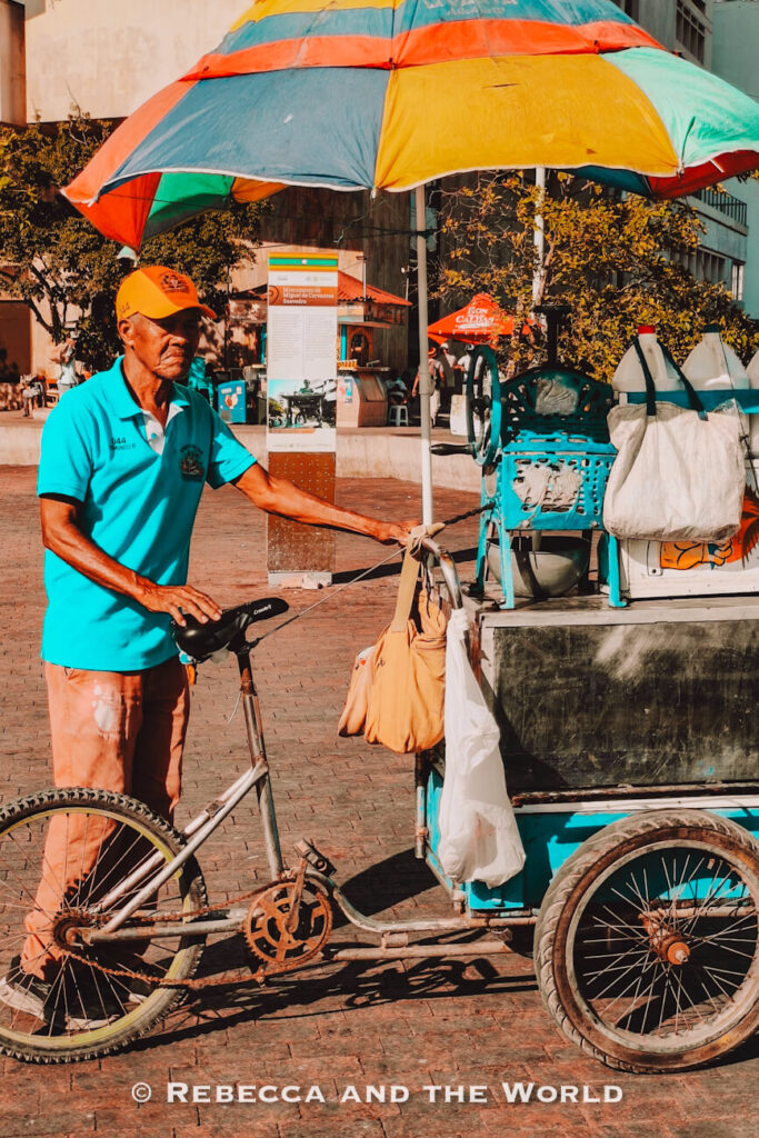 An older man in a turquoise polo shirt and orange cap stands beside a three-wheeled street cart under a large multicolored umbrella in a sunny plaza. Bags and containers hang from the cart, capturing the atmosphere of street food in Cartagena.