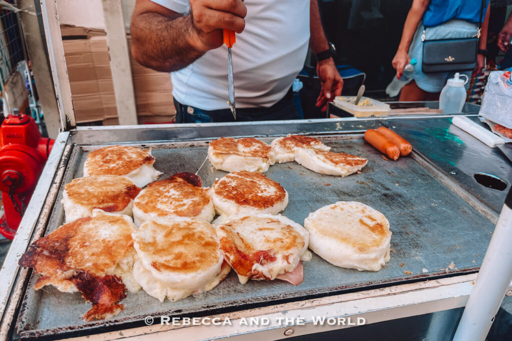 Several round stuffed arepas cook on a flat-top grill beside sausages while a vendor prepares food at a busy street stall during a Cartagena street food tour.