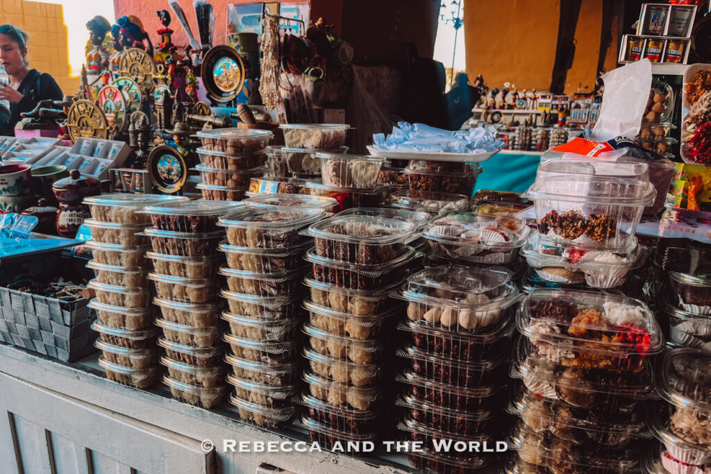 Clear plastic containers filled with packaged sweets and desserts are stacked on a market stall counter surrounded by souvenirs and local crafts in Cartagena.
