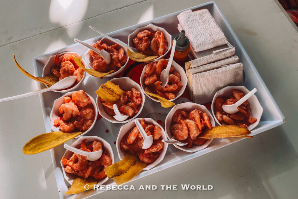 Small bowls of shrimp cocktail topped with plantain chips are arranged on a white serving tray with plastic spoons and napkins.