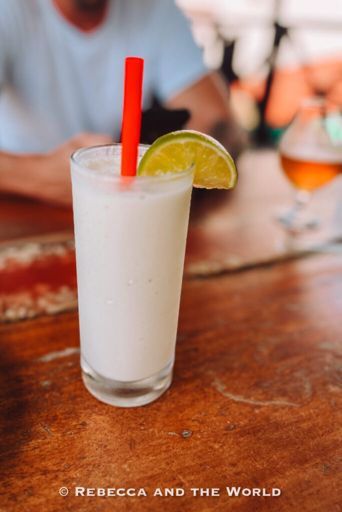 A creamy white drink garnished with a lime wedge and red straw sits on a wooden table with a blurred restaurant background, highlighting street food in Cartagena. This is limonada de coco, one of the best drinks in Colombia.